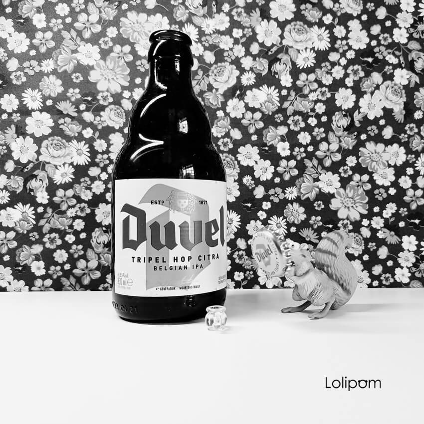 Photo noir et blanc sur fond fleuri d'une bière Duvel et d'un écureuil en jouet tenant la capsule de la bière avec un verre de bière à ses cotés bière